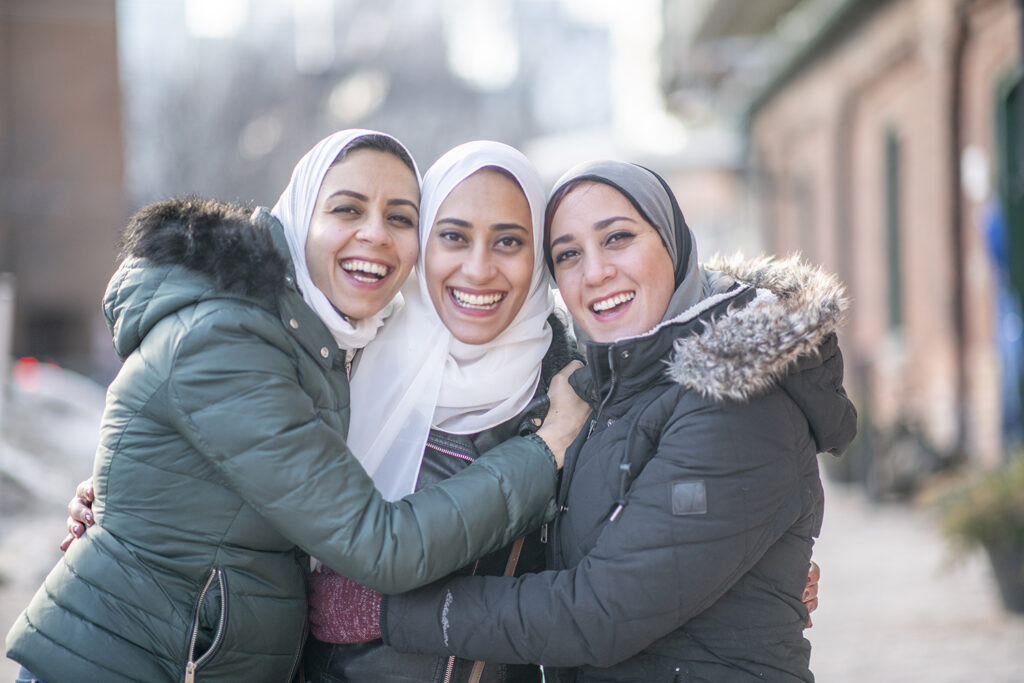 Three beautiful Muslim sisters standing outdoors on a winter day. They are embracing and hugging each other for a photo. They are happy and smiling at the camera.